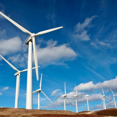 A row of white wind turbines on a hilly terrain. It's a sunny day with a few clouds in the sky.