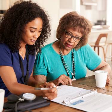 A young female nurse sits next to an older female patient at a wooden table and helps her fill out a form.