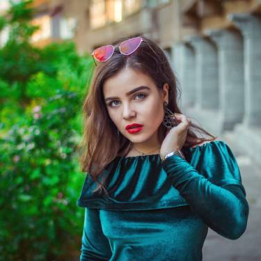 A model wearing red lipstick, sunglasses on her head, and a blue velvet top. She's pulling her hair behind her ear.