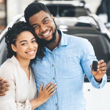 A couple smiling and showing off the key to their new car with the new white sedan in the background.