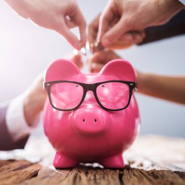 A close-up of a pink piggy bank wearing square black-rimmed glasses. There are four people putting money in the bank.