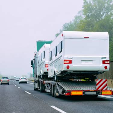 A large truck with a green cab transporting two white RVs on a highway. There are cars driving in front of the truck.